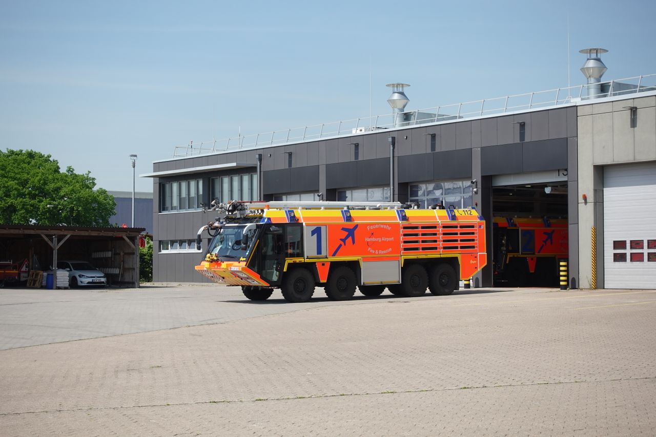 An airport fire truck parked outside a sleek fire station on a sunny day.